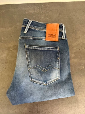 Replay Hyperflex blå jeans 32/32 - Snygga blå jeans från Replay Hyperflex med klassisk femficksdesign och ljusa slitningar på låren. Jeansen har raka ben och en skinnpatch med logga bak i midjan. Stretchigt material för extra komfort.