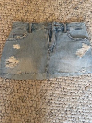 Ljusblå jeanskjol med slitningar - Trendig ljusblå jeanskjol i kort modell, från Hollister, i storlek W26, passar S. Midjemått ca 37