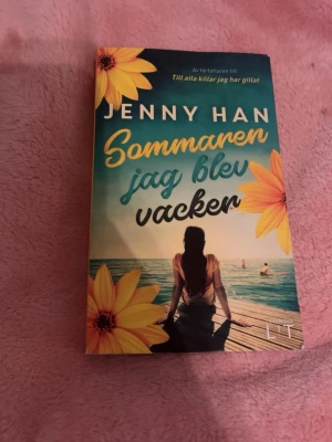 Sommaren jag blev vacker av Jenny Han - En ungdomsroman med färgstarkt omslag i blått, gult och grönt. Boken har mjukt pärmomslag och motiv av en tjej vid en brygga med stora gula blommor. Perfekt för dig som gillar romantik och sommarkänsla.
