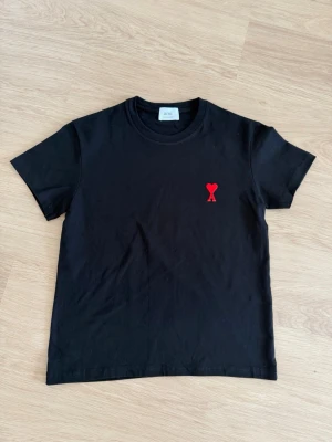 Svart t-shirt från Ami med rött hjärta - Snygg svart t-shirt från Ami med klassisk passform och rund hals. På bröstet finns ett broderat rött hjärta med bokstaven A, vilket ger en unik och stilren detalj. Tillverkad i mjuk bomull som känns skön mot huden. Perfekt för en clean och trendig look.