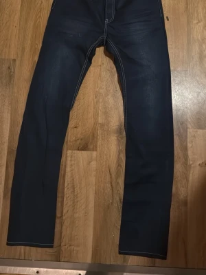 Mörkblå raka jeans med kontrastsömmar - Säljer ett par mörkblå jeans med raka ben och tydliga vita kontrastsömmar. Jeansen har klassisk femficksdesign och subtila slitningar framtill och baktill. Perfekta för dig som gillar en enkel men snygg stil.