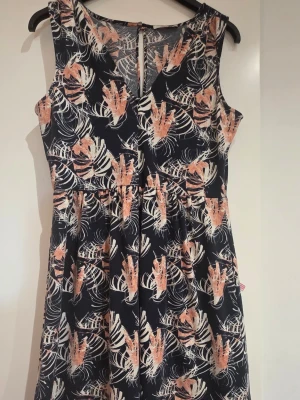 Blommig ärmlös klänning i marinblått - Snygg ärmlös klänning med marinblå bas och blommigt mönster i beige och orange toner. Klänningen har en markerad midja och lätt utställd kjol, perfekt för varma dagar. Tillverkad i mjukt och lätt material som känns skönt mot huden.