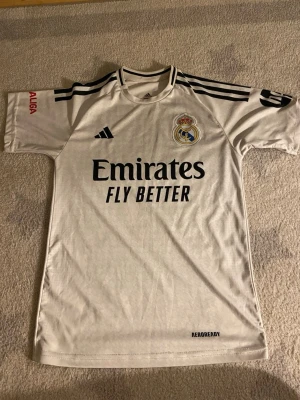 Real Madrid matchtröja Mbappé nr 9 - Vit Real Madrid matchtröja från Adidas med svarta detaljer, klubbmärke på bröstet och sponsortryck 'Emirates Fly Better'. Svarta ränder på axlarna och 'Mbappé 9' tryckt på ryggen. Tillverkad i lätt och ventilerande material.