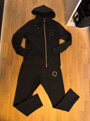 Svart loungewear-set från BOSS - Mörkblått loungewear-set från BOSS med dragkedja på huvtröjan och matchande byxor. Setet har diskret BOSS-logga på både tröja och byxa. Mjukt material och avslappnad passform, perfekt för chill dagar hemma.