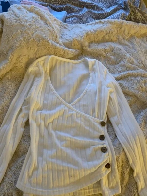 Vit omlottopp med knappar - Supersnygg vit omlottopp med ribbad struktur och tre dekorativa knappar i trä längs sidan. Toppen har lång ärm och v-ringning, perfekt att styla med jeans eller kjol för en trendig look.