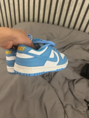 Nike Dunk Low blå och vita sneakers - Säljer ett par Nike Dunk Low sneakers i blått och vitt med gula Nike-loggor på hälen. Skorna har klassisk låg siluett, blå yttersula och vita paneler i läder. Perfekta för dig som gillar streetwear och vill sticka ut med färg. Har bara använts 3 gånger 
