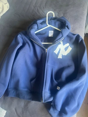 Blå zip-hoodie med vit logga - Blå new era hoodie med dragkedja från akimbo club Stor vit logga på bröstet, huva och känguruficka framtill. Mjuk bomullsblandning och ribbade muddar vid ärmslut och nederkant. Riktigt snygg hoodie tjock material,hög kvalité och sjukt nice fit. Tyvärr för liten för mig och skulle säga att den passar xs och S eftersom den är lite kortare. Helt ny aldrig använd eftersom jag beställde fel storlek. Priset kan diskuteras 