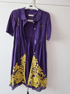Lila klänning med gul brodyr - Unik lila klänning från Odd Molly med gul broderad detalj nertill och dekorativa knappar framtill och på ärmarna. Klänningen har korta ärmar, klassisk krage och ett fint fall. Perfekt för dig som vill sticka ut med färg och mönster.