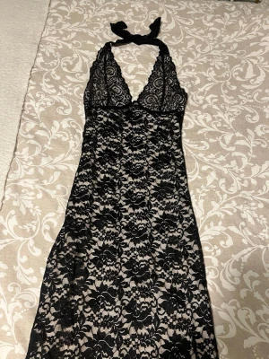 Svart spets klänning med halterneck - Klänning i svart spets med nude/beige foder och halterneck med knyt i nacken. Djup v-ringning och stretchigt material i polyester och elastan ger en bekväm och feminin känsla. Perfekt för dig som gillar eleganta detaljer.