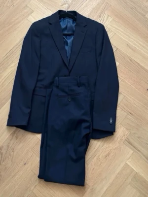 Mörkblå kostym - Snygga svarta kostymbyxor med rak siluett och klassisk design. Byxorna har knapp och dragkedja framtill samt fickor på sidorna. Perfekta för dig som gillar en stilren look och vill ha ett par byxor som funkar till skjorta eller kavaj. Materialet är troligtvis ull eller polyester. Både kavaj och byxor ingår. Dm mig för mer bilder eller frågor 