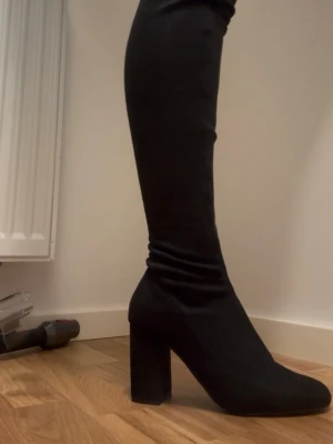 Svarta overknee stövlar med klack - Säljer ett par svarta overknee stövlar med hög blockklack och rund tå. Stövlarna är i stretchigt tyg som sitter tight längs benet och har en stilren, minimalistisk look. Perfekta för att lyfta vilken outfit som helst.