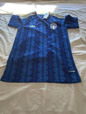 Blå Sverige fotbollströja Adidas - Säljer en blå fotbollströja från Adidas med Sveriges landslagsemblem på bröstet. Tröjan har gula ränder på axlarna, mönstrad design i olika blå nyanser och är tillverkad i lätt och ventilerande Climalite-material. Perfekt för träning eller match.