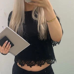 Svart croppad blus med spetsdetaljer - Svart croppad blus med fina spetsdetaljer längs ärmslut och nederkant. Blusen har korta ärmar och en lös passform som ger en avslappnad men ändå stilren look. Perfekt att matcha med höga jeans eller kjol.