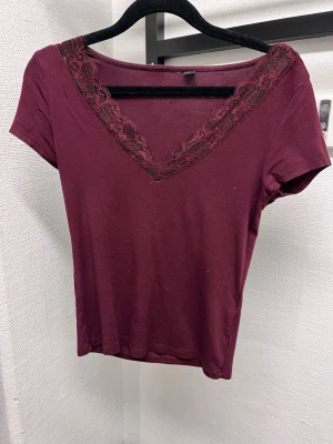 Vinröd topp med spetsdetalj - Vinröd topp med v-ringning och dekorativ spets längs halsringningen. Kortärmad och figurnära modell, perfekt att styla med jeans eller kjol. Mjuk och stretchig känsla, passar dig som gillar en enkel men snygg look. Är från brandy meville, knsppt använd men hsr lite smuts på sig, så om det önskas kan jag tvätta den innan jag skickar