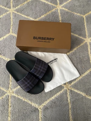 Burberry tofflor - Snygga svarta sandaler från Burberry med klassiskt rutmönster i mörkblått och grått på remmen. Tillverkade i slitstark syntet med präglad logga på fotbädden. Perfekta för en trendig och avslappnad stil.