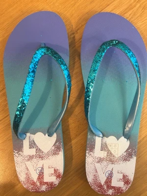 Blå glittriga flip-flops med LOVE-tryck - Snygga flip-flops i blått med glittriga remmar och texten LOVE på sulan. Remmarna är täckta av blått glitter och sulan har en snygg färgövergång från blått till vitt och rött. Perfekta för stranden eller sommardagar. Tillverkade i syntetmaterial.