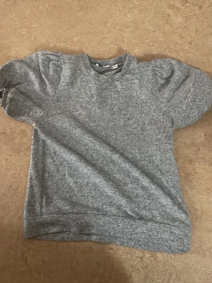 Grå topp med puffärmar - Grå melerad topp med rund hals och trendiga puffärmar. T-shirten har en avslappnad passform och är tillverkad i mjukt material, perfekt för en chill och stilren look.