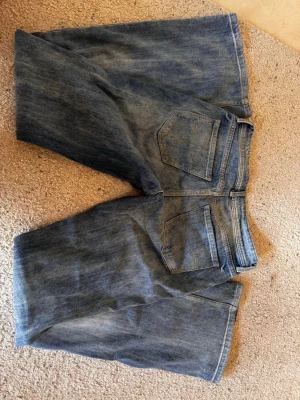 Blå bootcut jeans med slitningar - Snygga blå lågmidjade bootcut jeans i klassisk denim med lätt slitna detaljer och femficksmodell. Jeansen har vida ben, perfekta för en avslappnad och trendig look. 