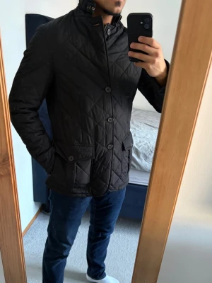 Barbour jacka - Elegant svart quiltad jacka från Barbour i storlek S med hög krage och spänne i halsen. Jackan har knapp- och dragkedjestängning, två framfickor med lock och en innerficka. Passar utmärkt nu till våren! Inga defekter! Bara att kontakta vid funderingar!🌟