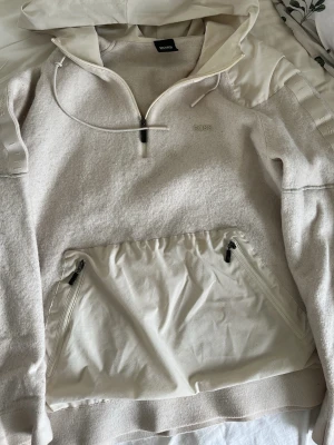 Beige hoodie från BOSS med dragkedja - Säljer en beige hoodie från BOSS med halv dragkedja framtill och stor huva. Hoodien har detaljer i nylon på axlar och ficka, samt två dragkedjeförsedda fickor framtill. Materialmix av mjuk bomull och nylon ger en modern look. Säljer på grund av ska flytta kan inte ta med alla kläder 