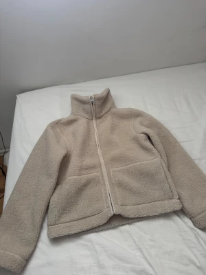 Beige teddyjacka med dragkedja - Mysig beige teddyjacka med hög krage och hel dragkedja framtill. Jackan har två stora fickor på framsidan och är tillverkad i fluffigt teddy-material som håller dig varm. Perfekt för kyliga dagar och enkel att matcha med olika outfits.