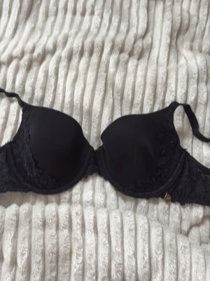 Svart push-up bh från Creem Luxury - Snygg svart push-up bh från Creem Luxury med spetsdetaljer på sidorna och en liten guldfärgad berlock framtill. Bh:n har vadderade kupor och justerbara axelband för bästa passform. Perfekt för dig som vill ha både komfort och stil.