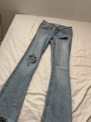 Ljusblå bootcut jeans med slitningar - Säljer ett par ljusblå bootcut jeans från Perfect Jeans med snygga slitningar på låret och knät. Jeansen har klassisk femficksdesign, låg midja och är tillverkade i denim. Perfekta för en avslappnad och trendig look.