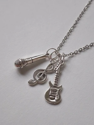 Halsband med musikinspirerade berlocker - Silvrigt halsband med tre berlocker: en mikrofon, en gitarr och en g-klav. Kedjan är tunn och smidig, och berlockerna har detaljerad design som passar dig som älskar musik och vill visa din stil. Perfekt accessoar för musikälskare.