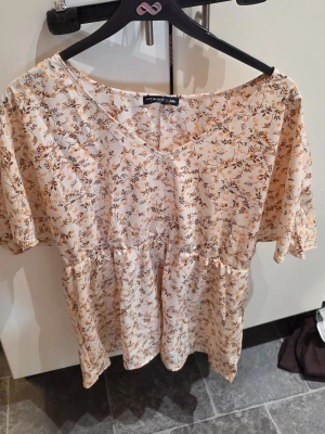 Blommig kortklänning från Vero Moda - Söt kortklänning från Vero Moda i ljusbeige med små blommor i orange och brunt. Klänningen har v-ringning, korta ärmar och ett avslappnat fall. Perfekt för varma dagar och enkel att styla med sneakers eller sandaler.