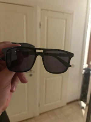 Gucci glasögon svara  - Snygga och stilrena solglasögon från Gucci i klassisk fyrkantig design. Modellen har en blank svart båge i acetat med mörktonade glas och diskret GUCCI-logga på sidorna, vilket ger en exklusiv men ändå clean look.
