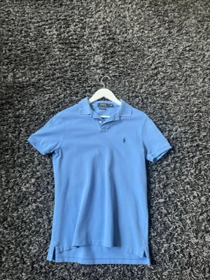 Blå pikétröja från Polo Ralph Lauren - Klassisk ljusblå pikétröja från Polo Ralph Lauren med korta ärmar och krage. Tröjan har knappar vid halsen och den ikoniska broderade loggan på bröstet. Perfekt för en avslappnad och stilren look. Skick 9/10. Skriv gärna frågor!