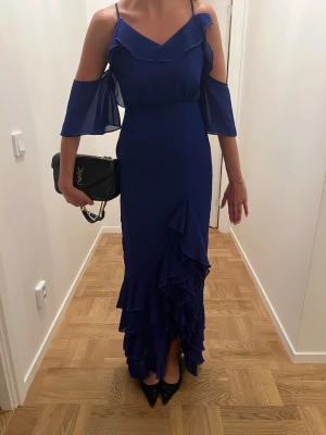 Marinblå långklänning med volanger - Snygg marinblå långklänning med volangdetaljer och offshoulder-ärmar. Klänningen har smala axelband och v-ringning, samt flera lager volanger nertill för en extra elegant look. Perfekt för dig som vill sticka ut med en stilren och trendig klänning. Perfekt inför balen, endast använd 1 gång