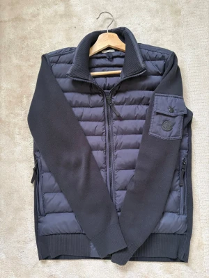 Mörkblå cardiganjacka från Moncler - Snygg mörkblå cardiganjacka från Moncler med quiltad front och stickade ärmar. Jackan har dragkedja, hög krage och en ficka med Moncler-logga på ärmen. Perfekt för lager-på-lager och streetstyle. Mycket mycket fint skick i princip oanvänt och jätte skonsamt använd. 