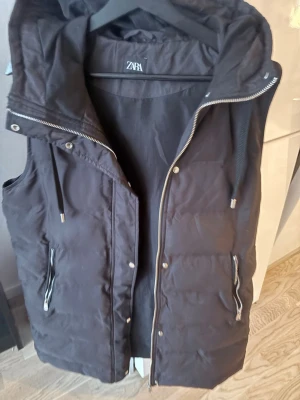 Svart dunväst med huva från Zara - Cool svart dunväst från Zara med dragkedja och två fickor med zip. Västen har en stor huva med snörning och quiltad design som ger en modern look. Perfekt för lager-på-lager och streetstyle. Materialet är syntet och fyllningen är dunliknande.