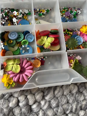 Pyssellåda med dekorationer scrapbooking  - En plastlåda fylld med blandade knappar i olika färger och storlekar, paljetter, glittriga dekorationer samt blommor i rosa, grönt och gult. Perfekt för kreativa projekt, scrapbooking och DIY-pyssel.