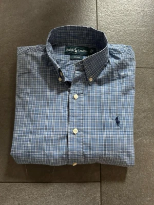 Blårutig skjorta från Ralph Lauren - Klassisk blårutig skjorta från Ralph Lauren med button-down krage och broderad logga på bröstet. Skjortan har långa ärmar och vita knappar framtill. Perfekt för en stilren och avslappnad look.