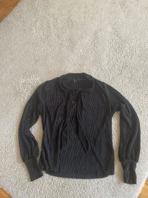 Svart blus med smala vita ränder - Snygg svart blus från Vera Moda  med smala vita ränder och knytband vid halsen. Blusen har långa ärmar med breda muddar och en rak passform. Perfekt för en trendig och stilren look.