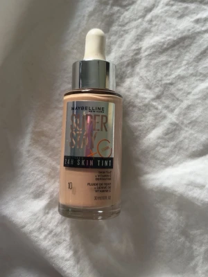 Maybelline Super Stay 24H Skin Tint 10 - Maybelline Super Stay 24H Skin Tint i nyansen 10. Flytande foundation med pipett, ger en naturlig finish och innehåller vitamin C. Kommer i en glasflaska med silverfärgat lock. Perfekt för dig som vill ha en lätt täckning och glow. Använd en gång. 