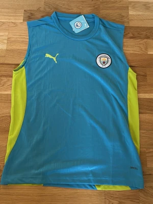 Manchester City ärmlös träningströja - Snygg ärmlös träningströja från Puma i Manchester Citys färger. Tröjan är blå med gula sidopaneler, klubbmärke på bröstet och Puma-logga. Tillverkad i lätt och ventilerande material, perfekt för intensiva träningspass.
