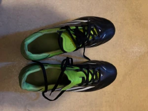 Svarta och gröna Adidas fotbollsskor - Säljer ett par snygga fotbollsskor från Adidas i svart med gröna och vita detaljer. Skorna har klassiska tre ränder på sidan, snörning och dobbar för spel på gräs. Insidan är grön och ovandelen är i syntetmaterial för bra bollkontroll.