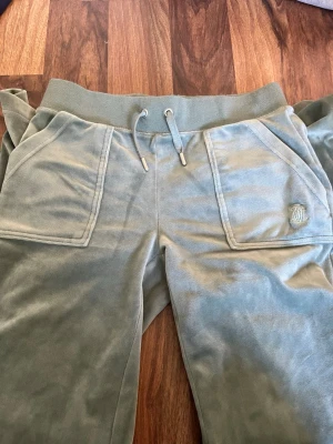 Gröna mjukisbyxor från Juicy Couture - Snygga gröna mjukisbyxor från Juicy Couture i mjuk velour. Byxorna har bred resår i midjan med dragsko, stora fickor fram och bakfickor med knappar. Ikonisk Juicy Couture-logga på ena fickan framtill. Perfekta för chill dagar, använd 1 gång då jag inte gillar färgen.