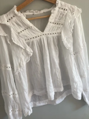 Vit romantisk blus med volanger - Superfin vit blus med volangdetaljer och broderade hål längs ärmar och bröst. Blusen har v-ringning, rynkade partier och långa ärmar med resår vid ärmslut. Perfekt för en söt och trendig look. Materialet känns lätt och luftigt, troligen bomull.