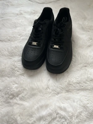 Nike Air Force 1 helsvarta sneakers - Säljer ett par helsvarta Nike Air Force 1 sneakers i klassisk low-modell. Skorna är i läder med perforerad tåbox, svarta skosnören och metall-detalj framtill. Swoosh-loggan syns på sidorna och sulan är i matchande svart. Perfekta för en clean och stilren look.