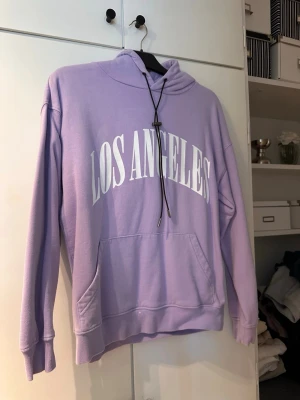 Lila hoodie med tryck 'Los Angeles' - Lila hoodie med vit text 'Los Angeles' på bröstet. Klassisk känguruficka framtill, huva med svarta dragsnören och ribbade muddar. Perfekt för en avslappnad streetstyle-look. Materialet känns mjukt och bekvämt.