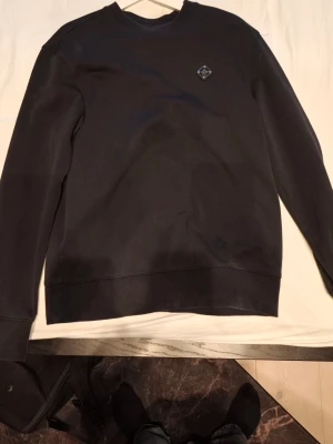 Svart sweatshirt från J.Lindeberg - En stilren svart sweatshirt från J.lindeberg med rund halsringning och diskret logomärke på bröstet. Tröjan har långa ärmar och ribbade muddar vid ärmslut och nederkant. Perfekt för en avslappnad och trendig look.