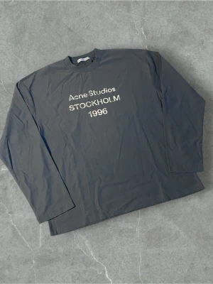 Blå långärmad topp Acne Studios | M - Svart långärmad topp från Acne Studios med vit text 'Acne Studios STOCKHOLM 1996' på bröstet. Rak passform och klassisk rund halsringning. Perfekt för en stilren och enkel look.
