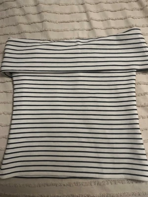 Randig offshoulder topp från Vila - Snygg vit offshoulder topp från Vila med svarta horisontella ränder. Toppen är figurnära och har en bred kant upptill som vilar över axlarna. Perfekt för dig som gillar en stilren och trendig look.