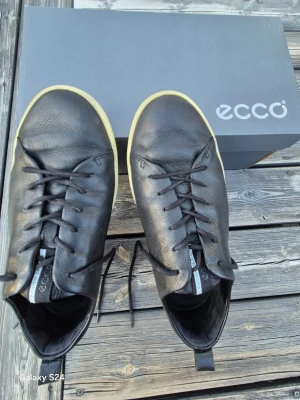 Svarta ECCO sneakers i läder - Snygga svarta sneakers från ECCO i mjukt läder med vita sulor och svarta skosnören. Klassisk låg modell med diskret ECCO-logga på plösen och dragflik bak. Perfekta för en stilren och avslappnad look.