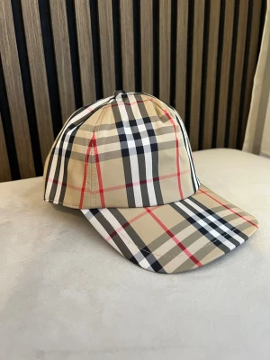 BBkeps med klassiskt rutmönster - Snygg keps från Burberry med det ikoniska beigea rutmönstret i svart, vitt och rött. Kepsen har böjd skärm och justerbar snapback baktill. Märkespatch med 'Burberry London England' på baksidan. Perfekt accessoar för en trendig look.