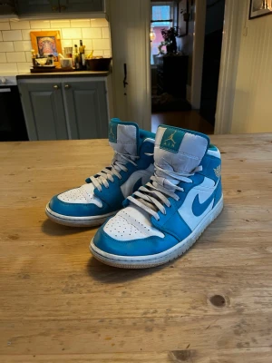 Nike Air Jordan 1 High OG blå/vit - Säljer ett par Nike Air Jordan 1 High OG i blått och vitt. Klassisk high-top siluett med vit bas, blå overlays och vit swoosh. Skorna har perforerad tåbox, blått Air Jordan Wings-märke och vit/blå plös med Air-logga. Ovandel i läder och klassisk yttersula.